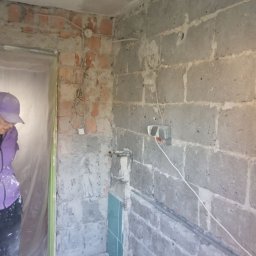 K & J Remont - Niedokończona łazienka z odsłoniętymi cegłami, blokami betonowymi, wystającymi przewodami, fragmentem niebieskich płytek i osobą w fioletowej bluzie i czapce z daszkiem w rogu kadru.
