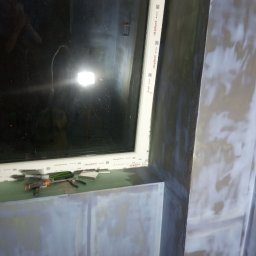 K & J Remont - Przygotowanie ściany do malowania: szpachlowana powierzchnia wokół okna z widocznymi narzędziami na parapecie.