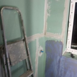 K & J Remont - Ściana w trakcie przygotowania do prac wykończeniowych w pomieszczeniu z oknem, widoczna drabina malarska, fragment hydroizolacji i niebieski element instalacji.