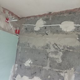 K & J Remont - Ściana z surowych betonowych bloków w trakcie przygotowywania do dalszych prac wykończeniowych, widoczne wyprowadzone przewody elektryczne w puszkach, częściowo zaszpachlowane bruzdy po przewodach.