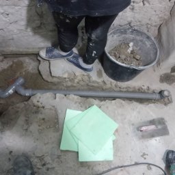 K & J Remont - Instalacja odpływu kanalizacyjnego w surowym wnętrzu, widoczne rury PCV, wiadro z gruzem, szpachelka i zielone płyty izolacyjne, osoba w roboczych ubraniach i tenisówkach w tle.