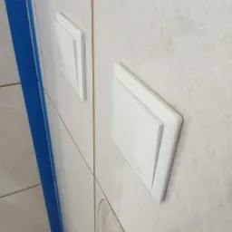 Dwa białe, prostokątne panele rewizyjne zamontowane na kremowych, błyszczących płytkach ściennych w łazience, z widocznym fragmentem niebieskiej listwy narożnej.