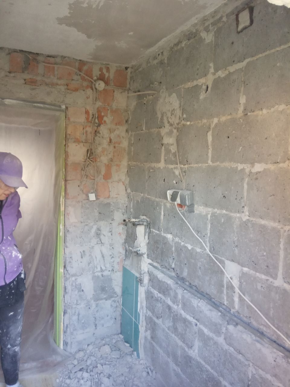 Niedokończona łazienka z odsłoniętymi cegłami, blokami betonowymi, wystającymi przewodami, fragmentem niebieskich płytek i osobą w fioletowej bluzie i czapce z daszkiem w rogu kadru.