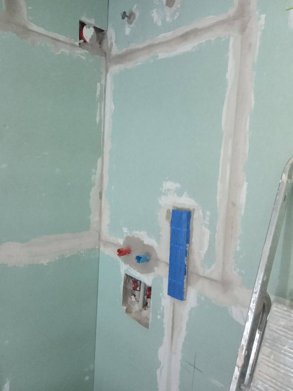 Ściana w trakcie przygotowania do wykończenia w łazience, z widocznymi rurami instalacji wodnej i elektrycznej, zabezpieczonymi niebieską taśmą, oraz częściowo zaszpachlowanymi łączeniami płyt...