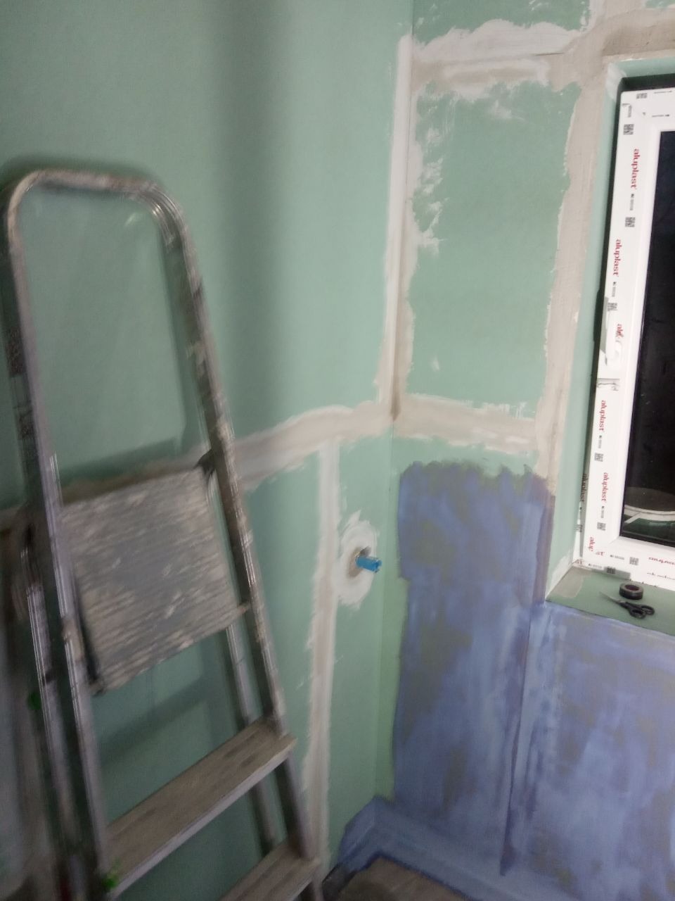 Ściana w trakcie przygotowania do prac wykończeniowych w pomieszczeniu z oknem, widoczna drabina malarska, fragment hydroizolacji i niebieski element instalacji.