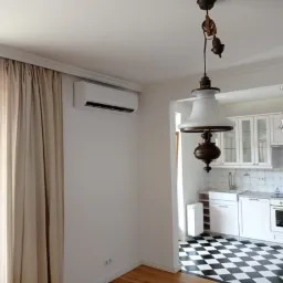 Biały klimatyzator ścienny zamontowany w jasnym pokoju z widokiem na kuchnię z czarno-białą podłogą w kratkę, obok okna z beżowymi zasłonami i ozdobnej lampy wiszącej