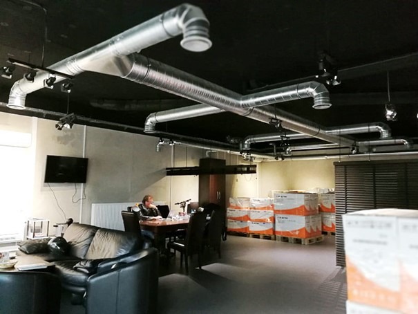 Biuro z widoczną instalacją wentylacyjną pod sufitem, oświetleniem punktowym, biurkiem, sofą i zapasem materiałów na paletach w tle.