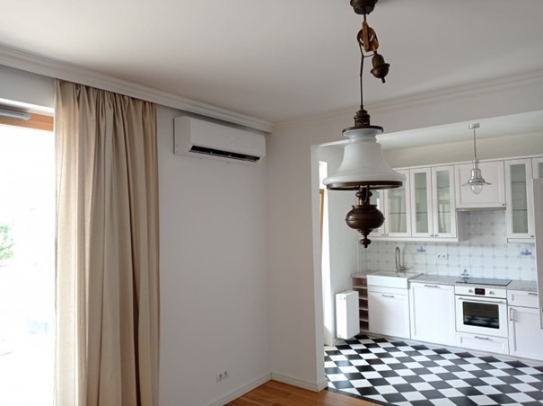 Biały klimatyzator ścienny zamontowany w jasnym pokoju z widokiem na kuchnię z czarno-białą podłogą w kratkę, obok okna z beżowymi zasłonami i ozdobnej lampy wiszącej