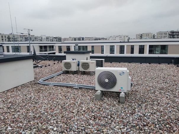 Instalacja klimatyzacji na dachu budynku mieszkalnego z widocznymi jednostkami zewnętrznymi i szarymi rurami odprowadzającymi, tło stanowi zabudowa miejska w pochmurny dzień.