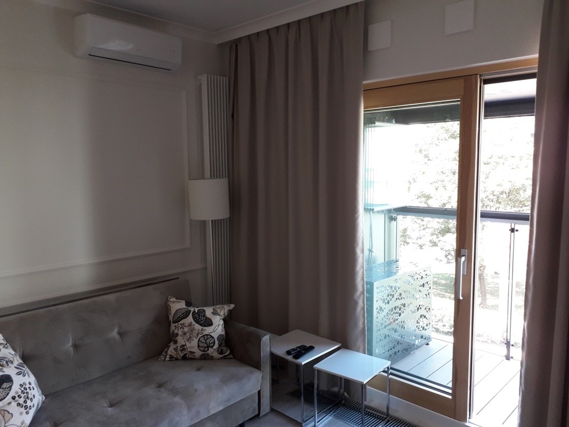 Klimatyzator ścienny zamontowany w pokoju dziennym z szarą sofą, lampą podłogową i wyjściem na balkon.