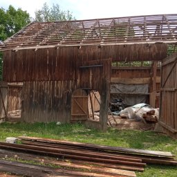 Łukasz Szymczyk - Częściowo rozebrany drewniany budynek gospodarczy z widoczną konstrukcją dachu, stertą desek na trawie i quadem wewnątrz.