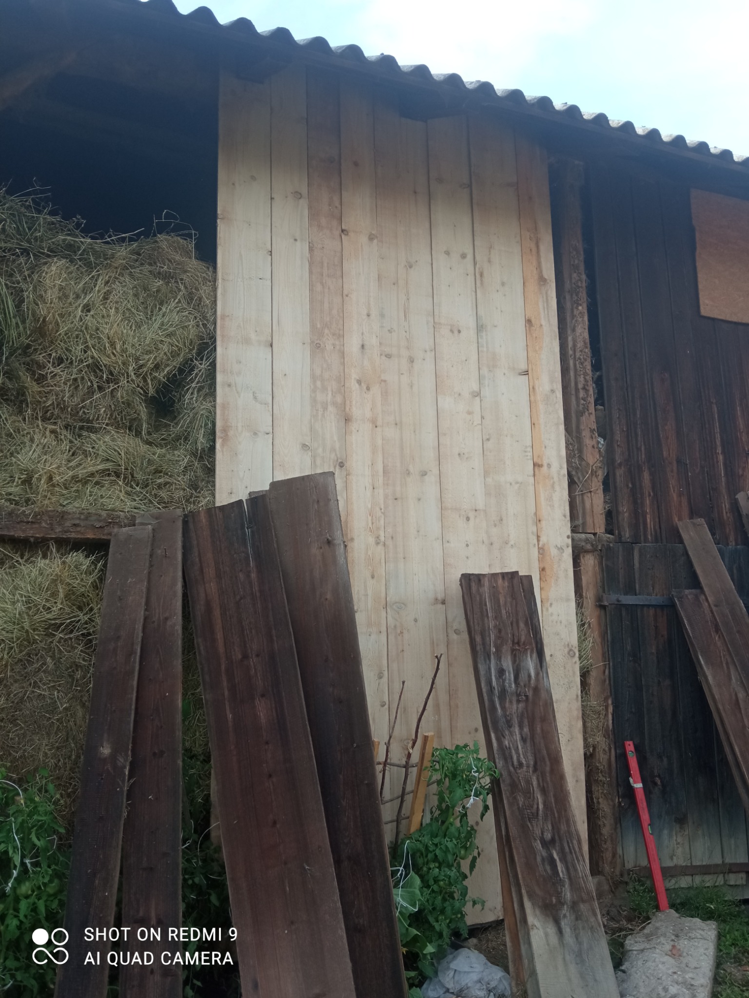 Drewniana ściana z desek w stodole, częściowo zasłaniająca siano, z ułożonymi deskami na ziemi i poziomnicą opartą o ścianę.