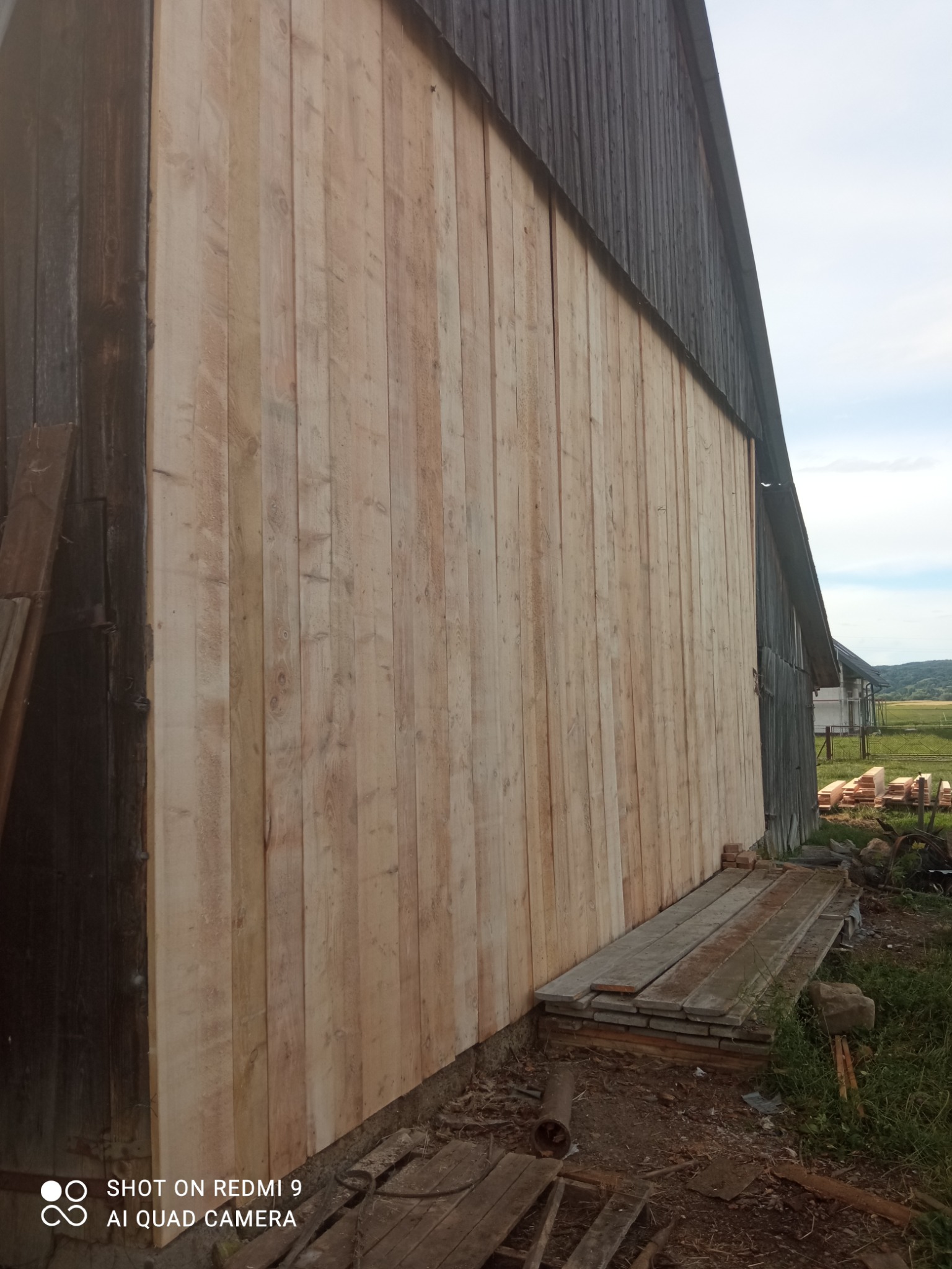 Fragment drewnianej ściany budynku w trakcie remontu, z widoczną nową, jasną boazerią kontrastującą ze starą, ciemną częścią konstrukcji, z podłożonymi płytami betonowymi.