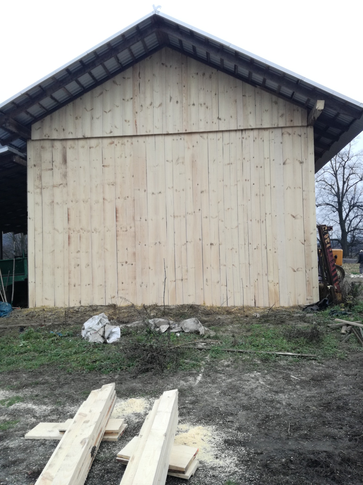 Nowa, jasna elewacja z desek na budynku gospodarczym, widoczny fragment dachu z blachy, teren wokół z resztkami trawy i błota, w tle drzewo bez liści i fragment traktora.