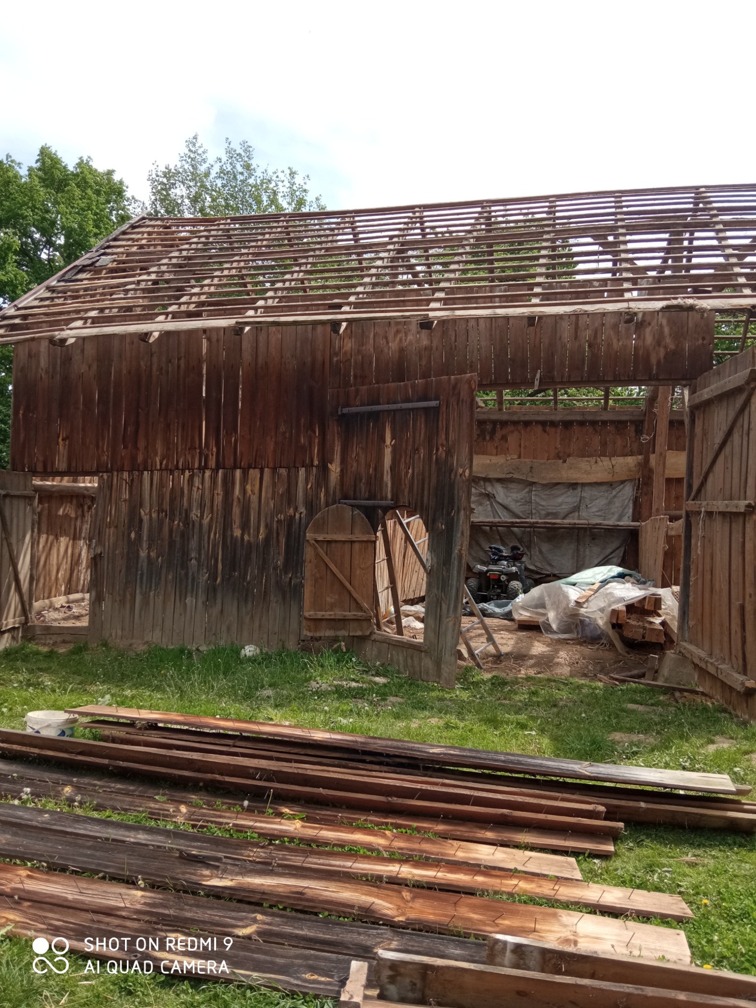 Częściowo rozebrany drewniany budynek gospodarczy z widoczną konstrukcją dachu, stertą desek na trawie i quadem wewnątrz.