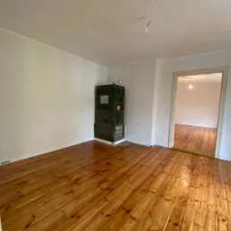 Pokój z wypolerowaną, lakierowaną podłogą z desek, ciemnozielonym piecem kaflowym w rogu i widokiem na kolejne pomieszczenie przez otwarte drzwi.