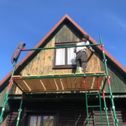 Mężczyzna maluje drewnianą elewację domu z rusztowania; widoczny fragment dachu, okno i wisząca bluza.