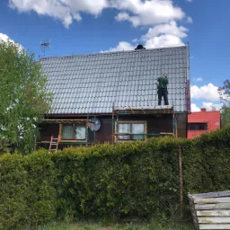 Pracownik w zielonej koszulce na rusztowaniu maluje fragment szarego dachu domu z ciemnego drewna, widok zza żywopłotu i metalowej siatki.