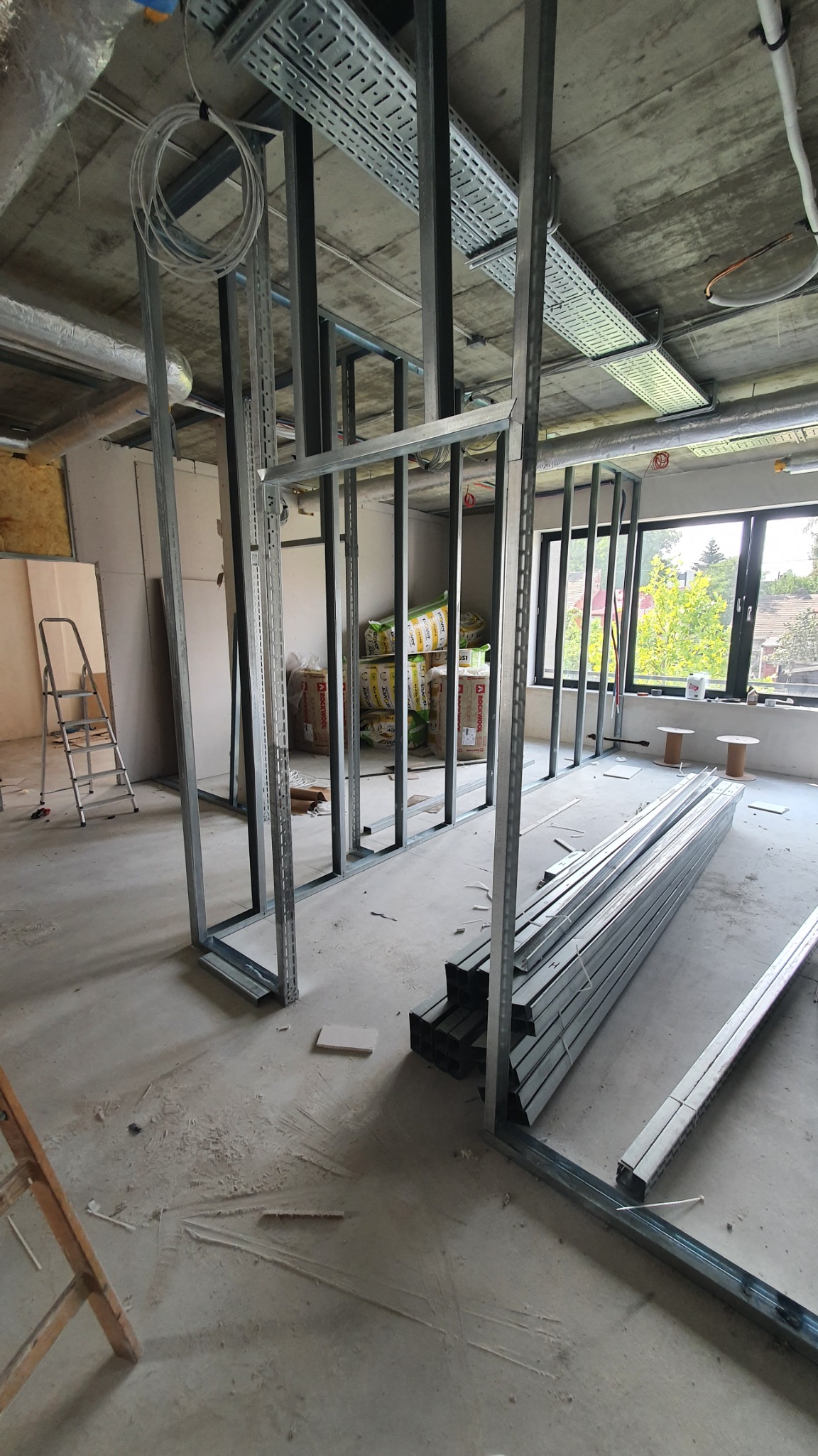 Metalowa konstrukcja ścian działowych w trakcie montażu, widoczne profile, przewody elektryczne pod sufitem, drabina i materiały izolacyjne w tle.