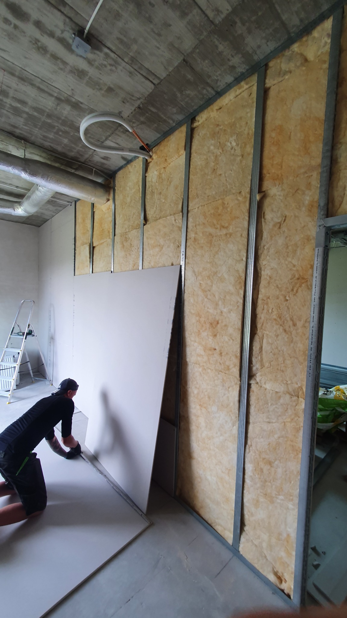 Pracownik docina dolną krawędź płyty gipsowo-kartonowej przy ścianie z izolacją z wełny mineralnej i metalowym stelażem, widoczna drabina w tle.