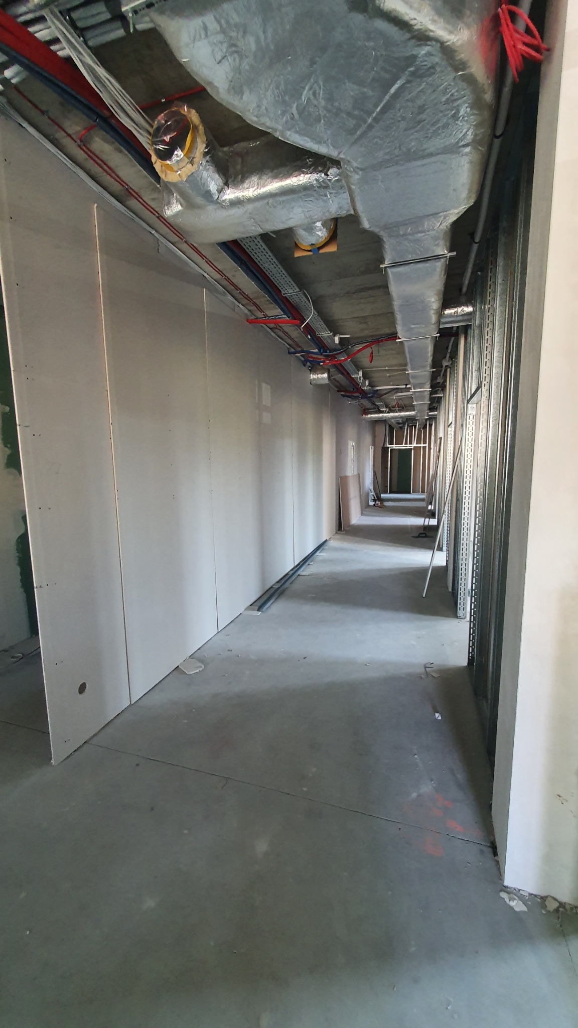 Korytarz w trakcie prac wykończeniowych, widoczne płyty gipsowo-kartonowe na ścianach i instalacje wentylacyjne pod sufitem.