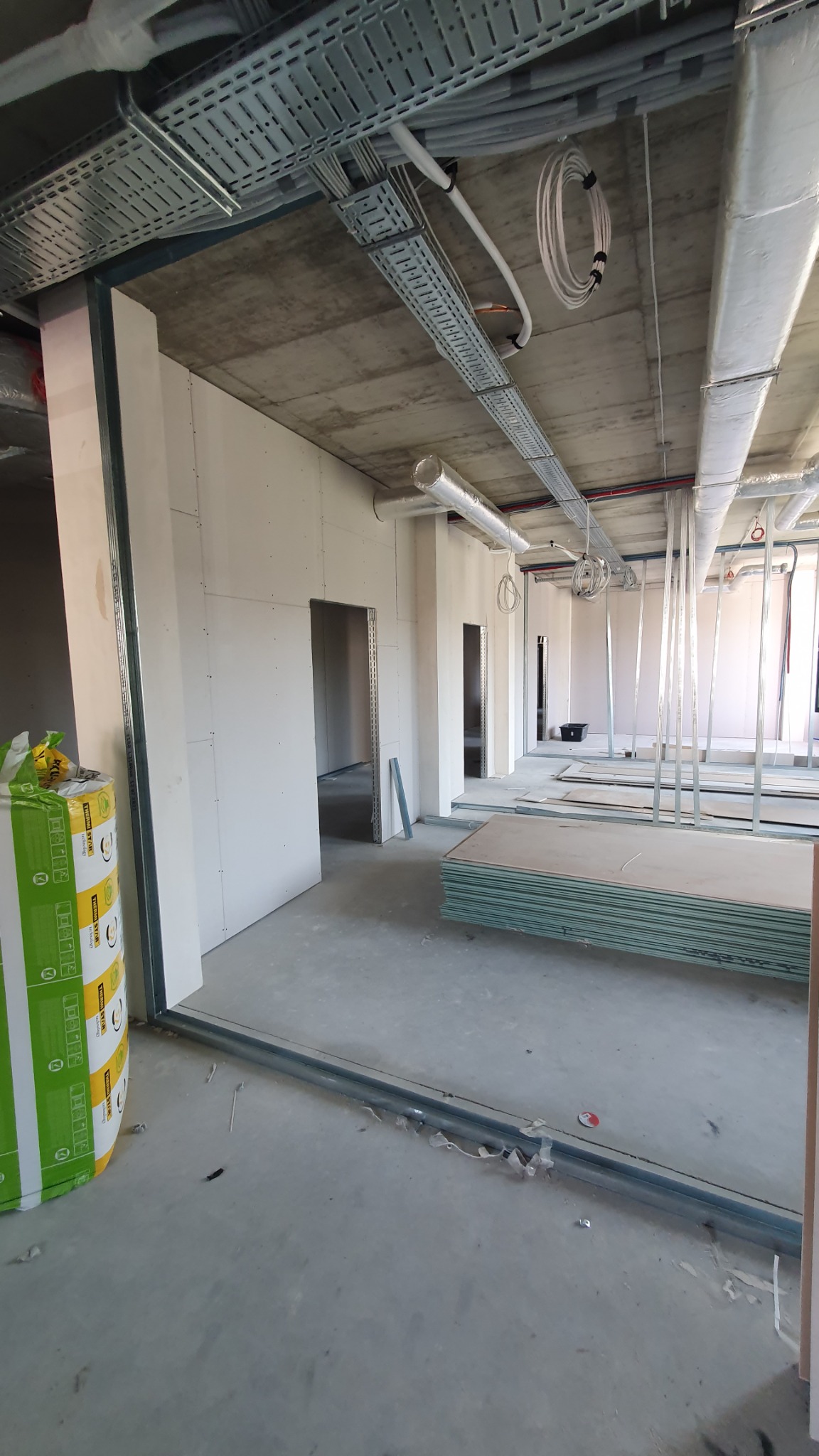 Wnętrze w trakcie wykańczania: widoczne profile metalowe, ułożone płyty gipsowo-kartonowe, odsłonięty betonowy sufit z instalacjami i otwarte przejścia.