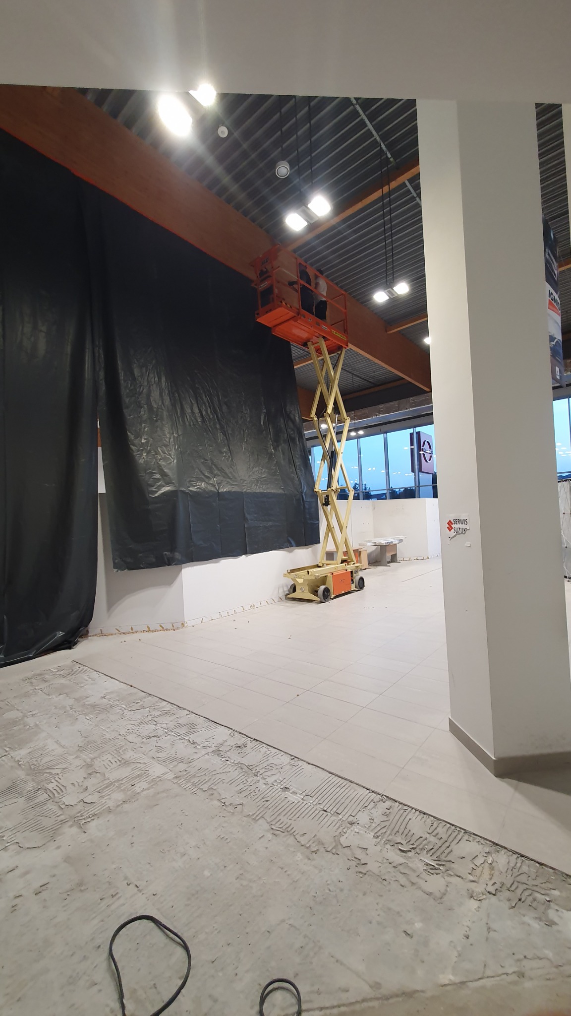 Wnętrze w trakcie prac wykończeniowych z podnośnikiem nożycowym na tle zasłoniętej ściany i fragmentu posadzki przed położeniem płytek.