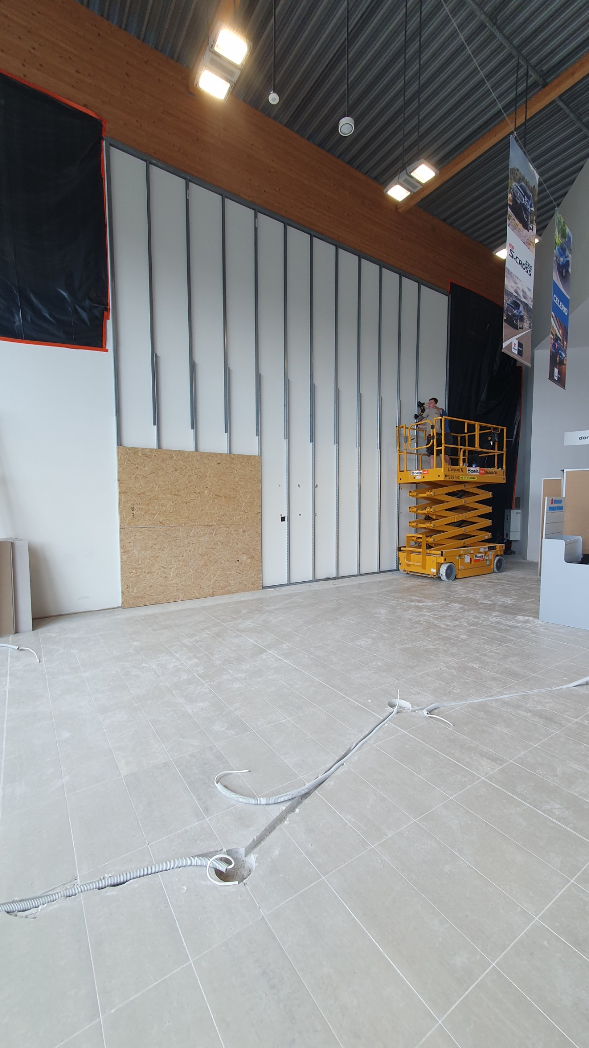 Montaż ścian działowych w nowym biurze: widoczne profile, płyty częściowo zamontowane, pracownik na podnośniku nożycowym, instalacja elektryczna w podłodze.