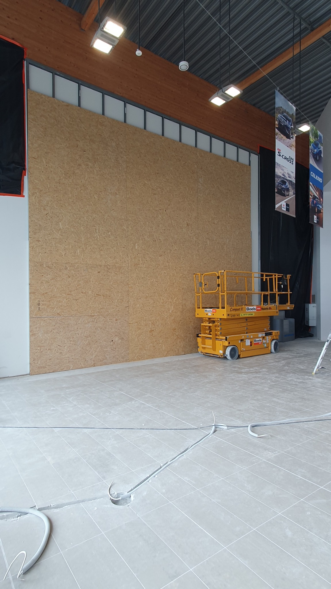 Wnętrze sklepu w trakcie prac wykończeniowych, widoczna ściana z płyty OSB, podnośnik nożycowy i wystające kable z podłogi.