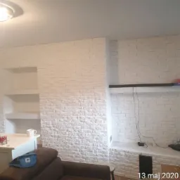 Biała ściana z cegły z wnękami w salonie w Andrychowie, na półkach książki i dekoracje.