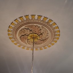 Pawel Plonka - Ozdobny, okrągły element dekoracyjny sufitu z motywem roślinnym w odcieniach beżu i żółci, z łańcuchem zwisającym od żyrandola kryształowego z zapalonymi żarówkami.