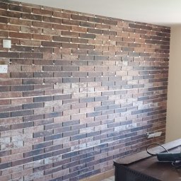 Pawel Plonka - Ściana z dekoracyjnej cegły w trakcie montażu, widoczne plastikowe krzyżyki dystansowe, drewniana podłoga i komoda w tle.