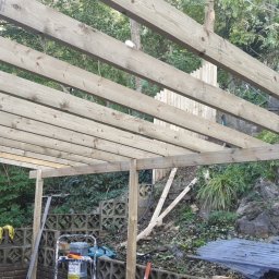 Pawel Plonka - Konstrukcja drewniana z belek stropowych w ogrodzie, widoczne narzędzia i materiały budowlane w tle roślinność i schody.