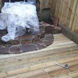 Pawel Plonka - Nowy drewniany taras z desek ułożonych równolegle, obok kamiennych schodów i muru, częściowo przykrytych folią ochronną, z narzędziem pomiarowym leżącym na deskach.