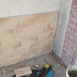 Pawel Plonka - Fragment ściany z częściowo ułożonymi panelami imitującymi kamień, widoczne narzędzia i materiały budowlane, w tle surowa ściana i narożnik z imitacją cegły.