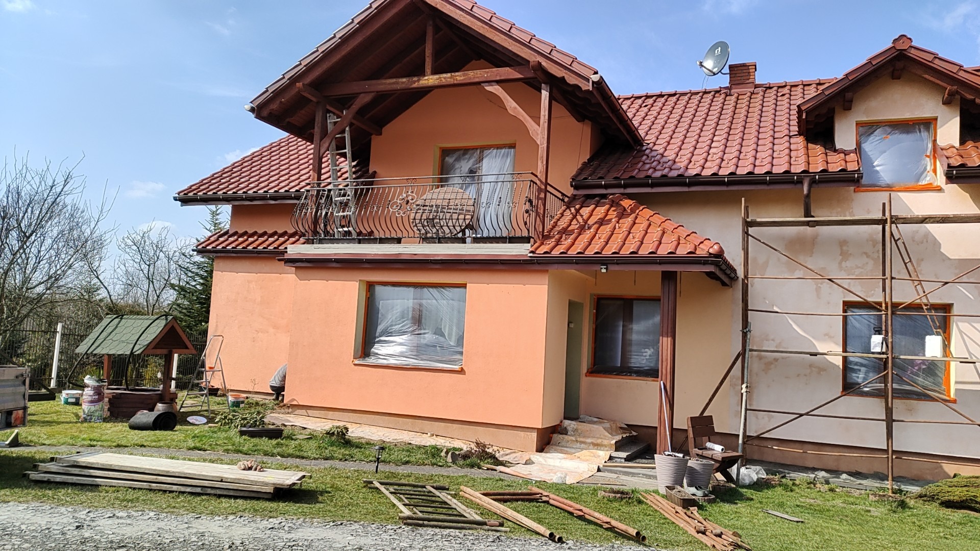 Dwukondygnacyjny dom jednorodzinny z balkonem i czerwoną dachówką, częściowo osłonięty rusztowaniem, z widocznymi śladami prac renowacyjnych elewacji, okna zabezpieczone folią, na trawniku leżą...