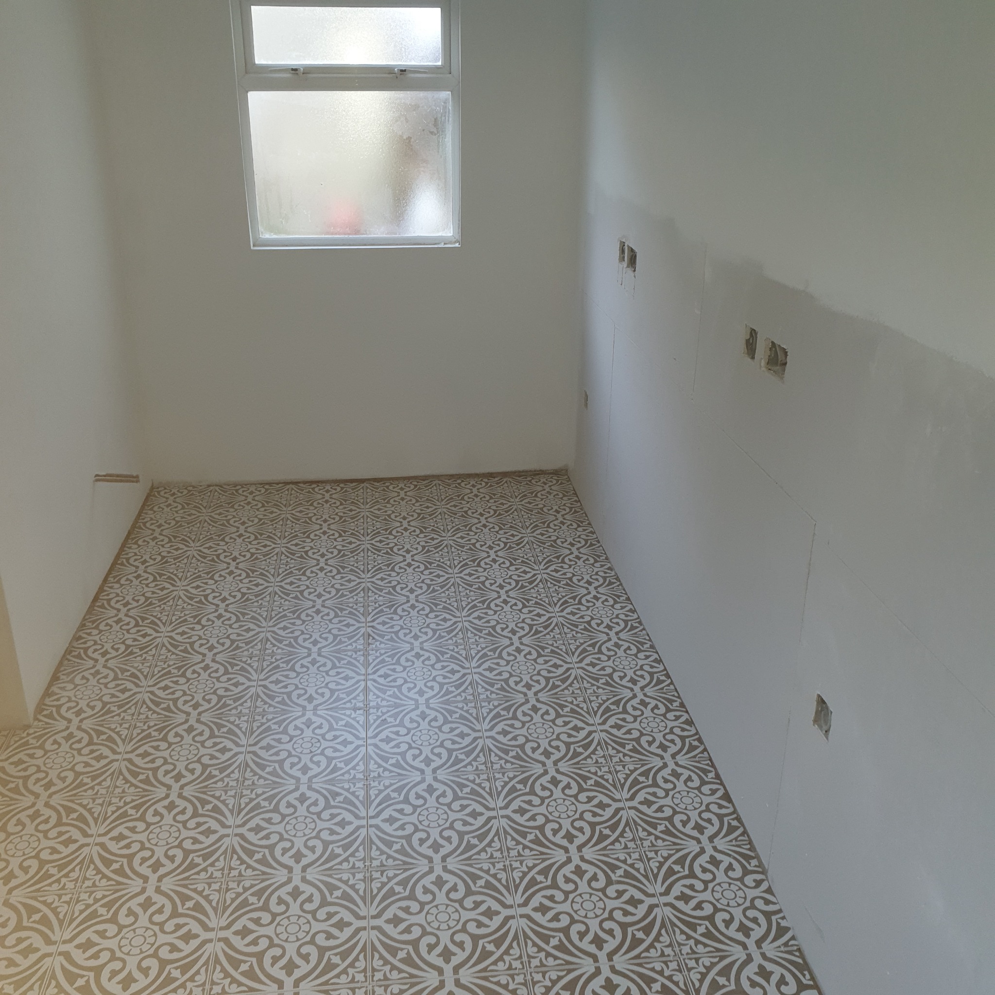 Wnętrze pomieszczenia po remoncie: wzorzysta, beżowo-biała podłoga z płytek, białe ściany z widocznymi fragmentami po szpachlowaniu i otworami na gniazdka, małe okno z matową szybą.
