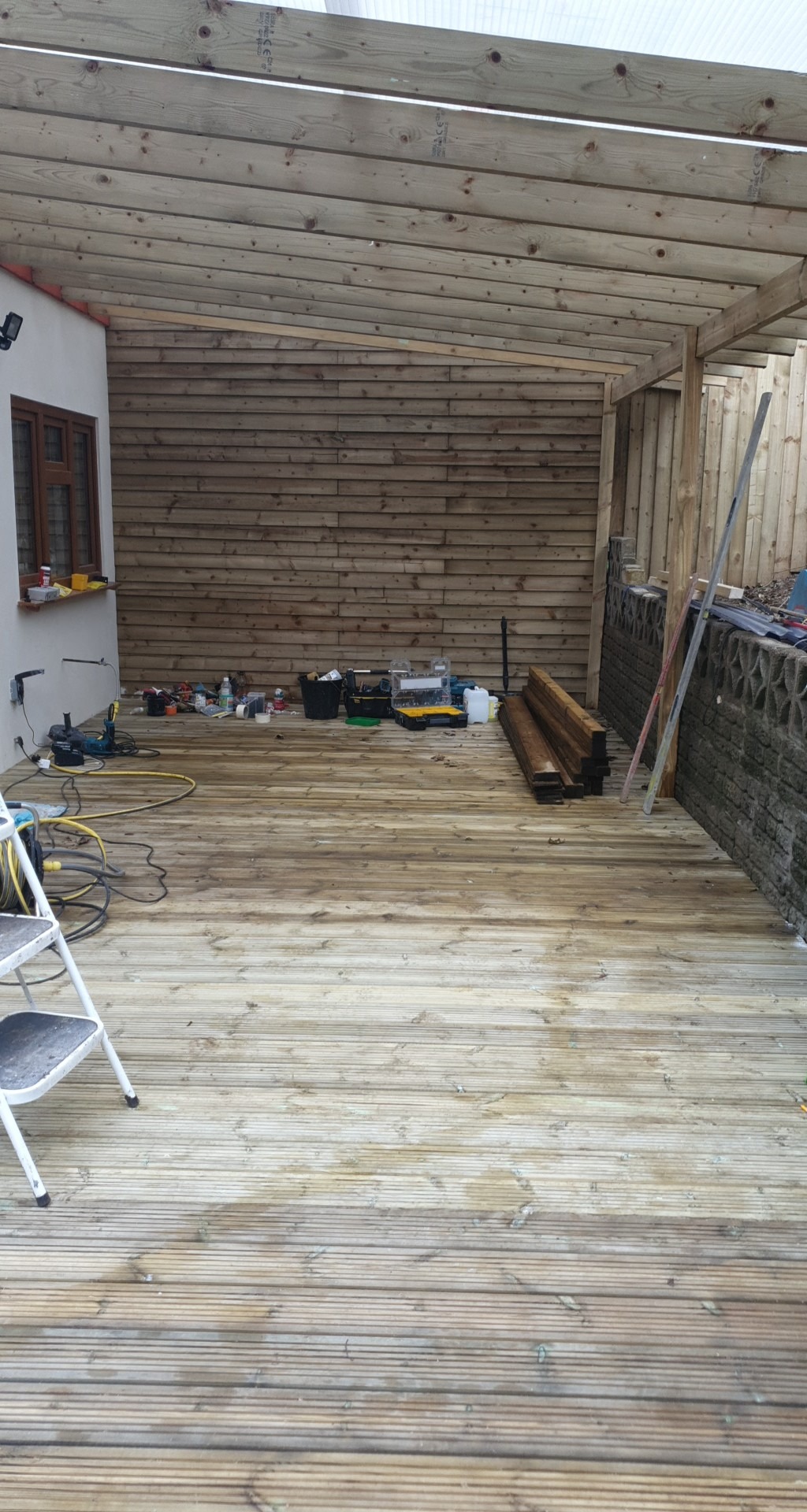 Drewniany taras w trakcie budowy z częściowo zadaszoną konstrukcją, widoczne narzędzia i materiały budowlane, schodki malarskie po lewej stronie, ściana z drewnianych paneli w tle.
