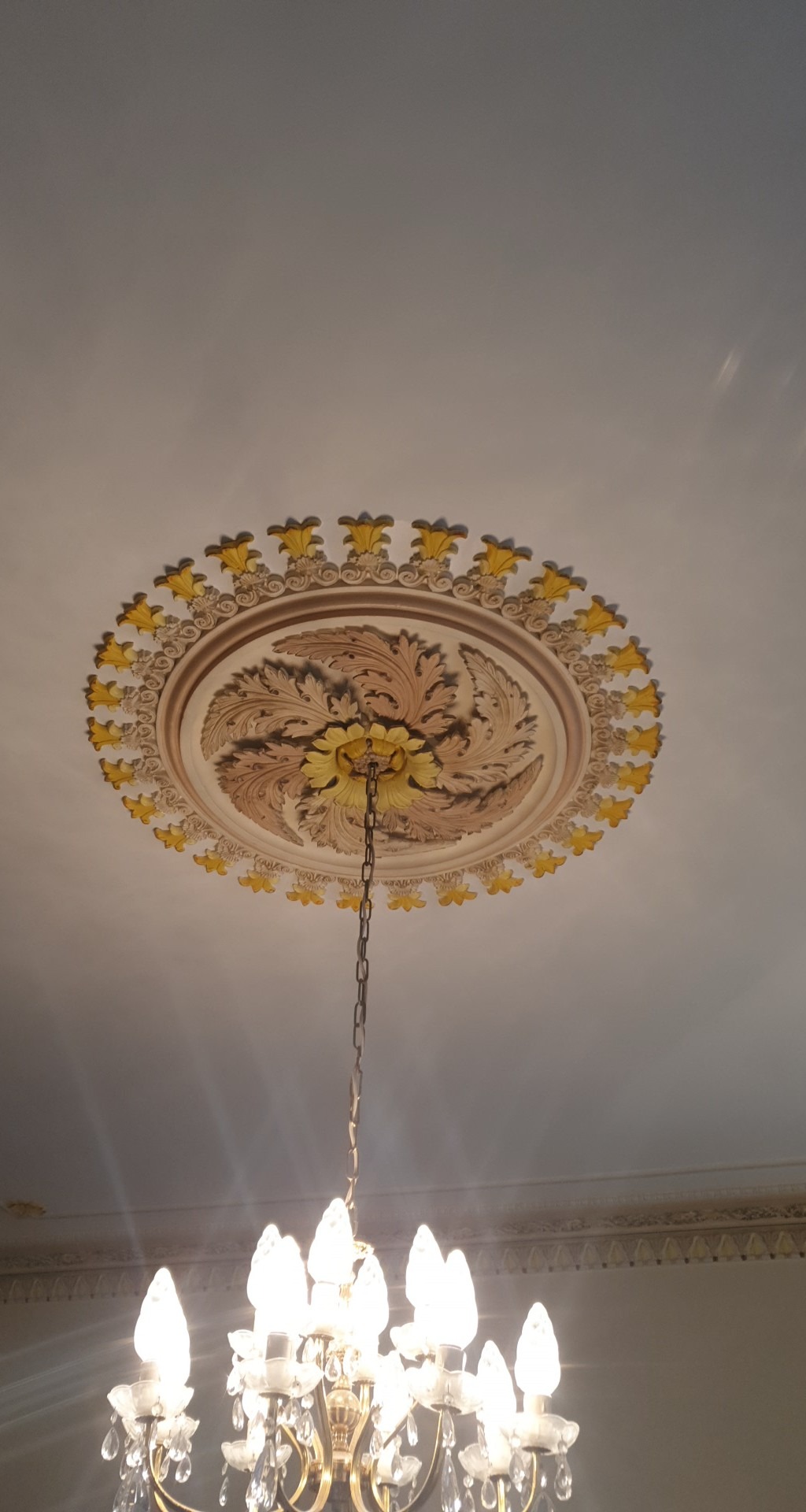 Ozdobny, okrągły element dekoracyjny sufitu z motywem roślinnym w odcieniach beżu i żółci, z łańcuchem zwisającym od żyrandola kryształowego z zapalonymi żarówkami.
