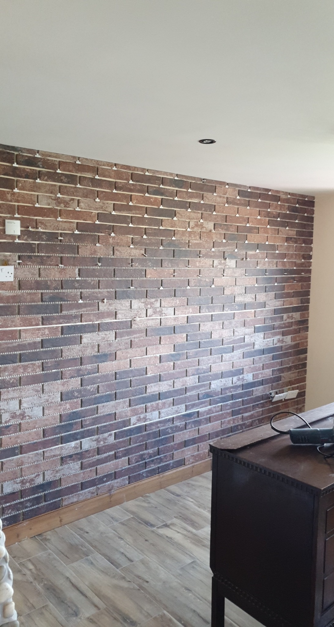 Ściana z dekoracyjnej cegły w trakcie montażu, widoczne plastikowe krzyżyki dystansowe, drewniana podłoga i komoda w tle.