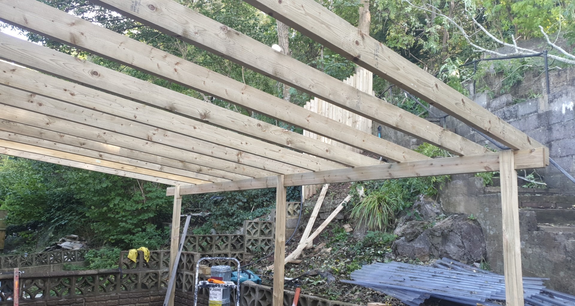 Konstrukcja drewniana z belek stropowych w ogrodzie, widoczne narzędzia i materiały budowlane w tle roślinność i schody.