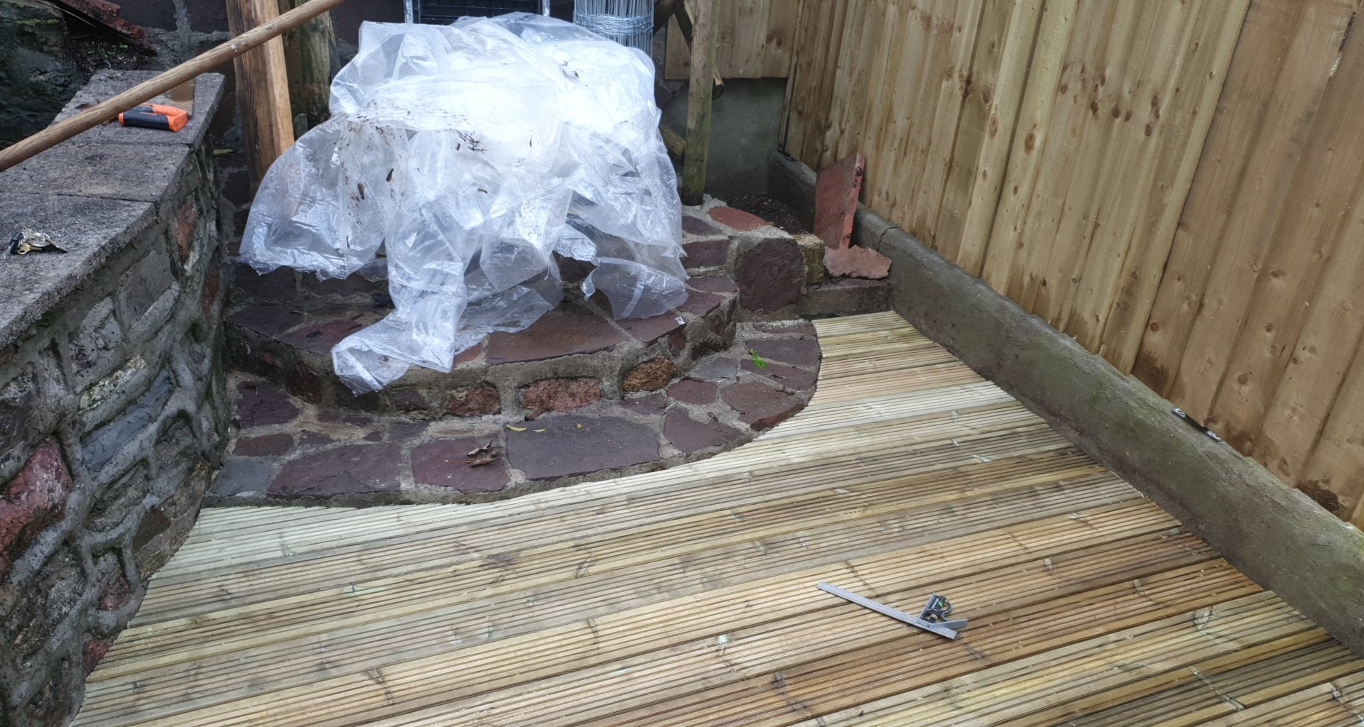 Nowy drewniany taras z desek ułożonych równolegle, obok kamiennych schodów i muru, częściowo przykrytych folią ochronną, z narzędziem pomiarowym leżącym na deskach.