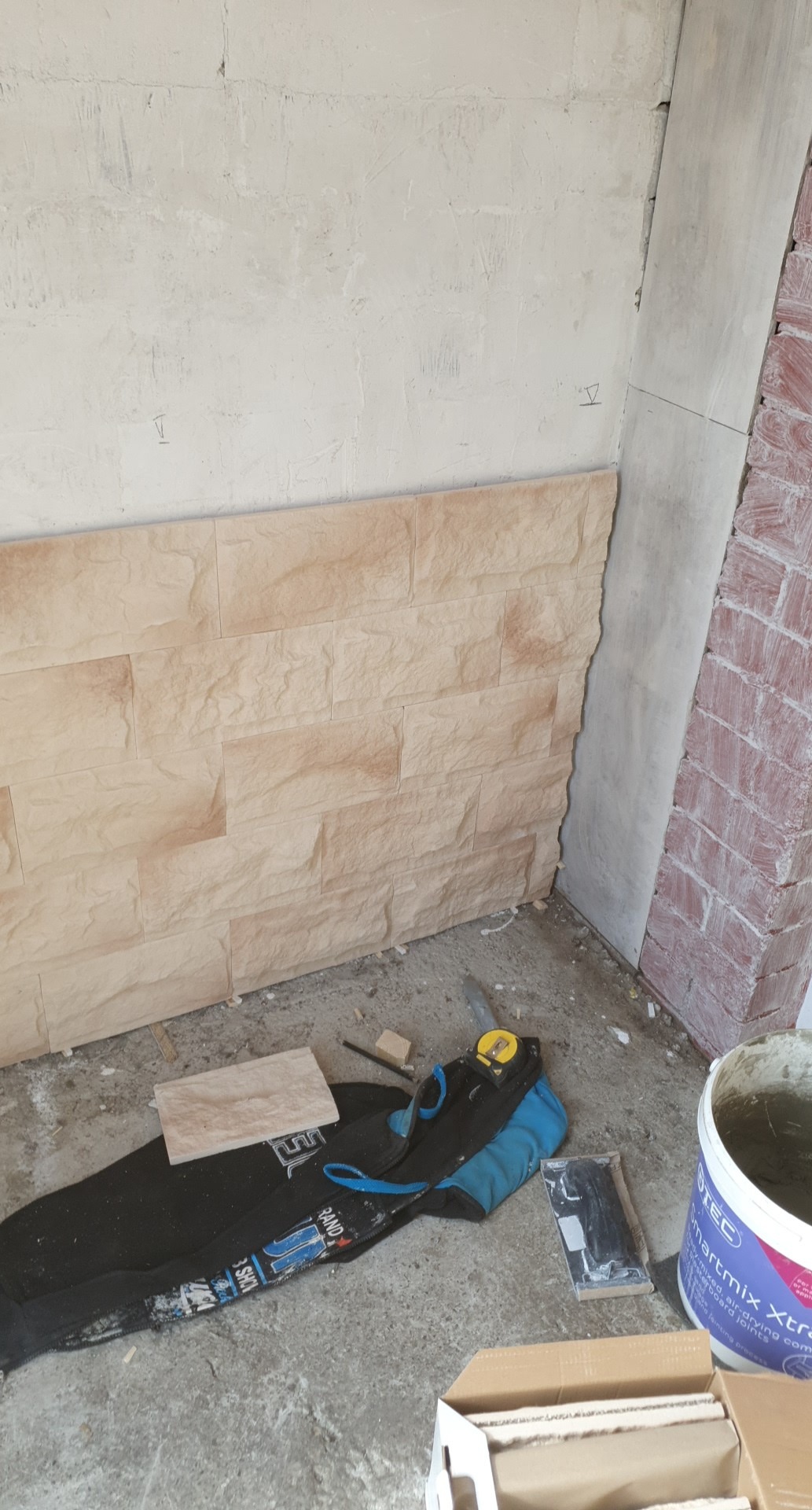 Fragment ściany z częściowo ułożonymi panelami imitującymi kamień, widoczne narzędzia i materiały budowlane, w tle surowa ściana i narożnik z imitacją cegły.