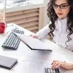 Kobieta w okularach, w białej koszuli, siedzi przy biurku, przegląda dokumenty i korzysta z kalkulatora. Na biurku komputer, klawiatura, alarm, notatnik i pojemnik z długopisami.