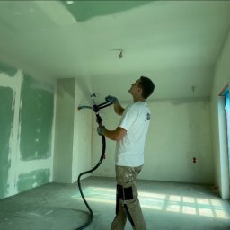 Busz Remonty - Pracownik w okularach ochronnych natryskuje gładź na sufit w pomieszczeniu z częściowo wykończonymi ścianami, używając profesjonalnego pistoletu natryskowego z długim wężem.