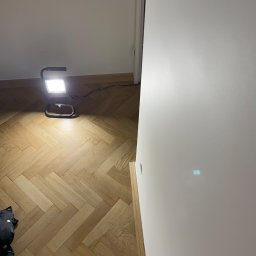 Busz Remonty - Świeżo pomalowana, biała ściana podświetlona lampą roboczą na tle parkietu ułożonego w jodełkę.