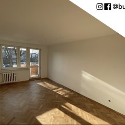 Busz Remonty - Pusty pokój po świeżym pomalowaniu na biało, parkiet w jodełkę, widoczne okno balkonowe i grzejnik, w prawym górnym rogu logo firmy.