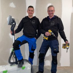 Busz Remonty - Dwóch mężczyzn w odzieży roboczej, prezentujących szlifierkę do gładzi i wkrętarkę na tle ściany po szpachlowaniu, z widocznymi łatami i nierównościami.