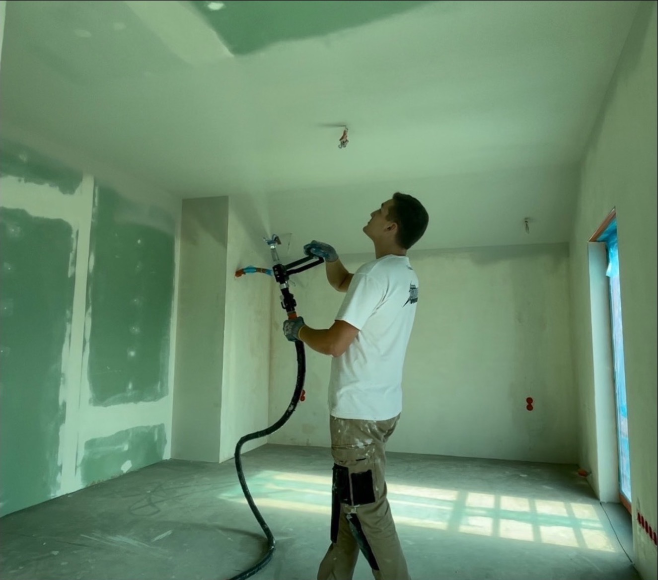 Pracownik w okularach ochronnych natryskuje gładź na sufit w pomieszczeniu z częściowo wykończonymi ścianami, używając profesjonalnego pistoletu natryskowego z długim wężem.