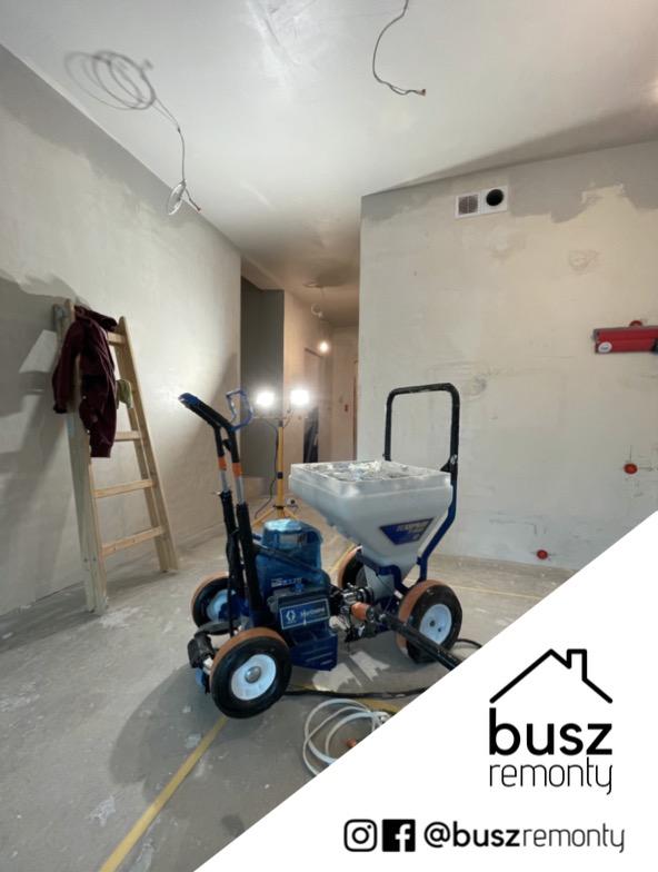 Urządzenie natryskowe do gładzi na tle wnętrza w trakcie prac wykończeniowych, widoczna drabina i niedokończone ściany, oświetlenie halogenowe. Logo firmy remontowej w rogu.
